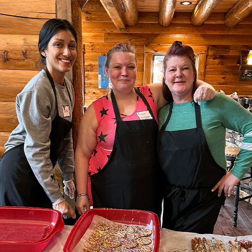 Three women wearing aprons gathered around desserts