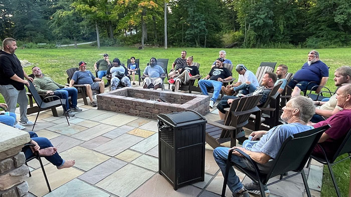 A single man, addressing a large group of men all gathered around a campfire.